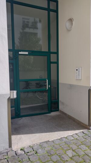 Foto der gläsernen Eingangstür, mit gründer Metallfassung. Rechts an der Wand befindet sich die Klingel.