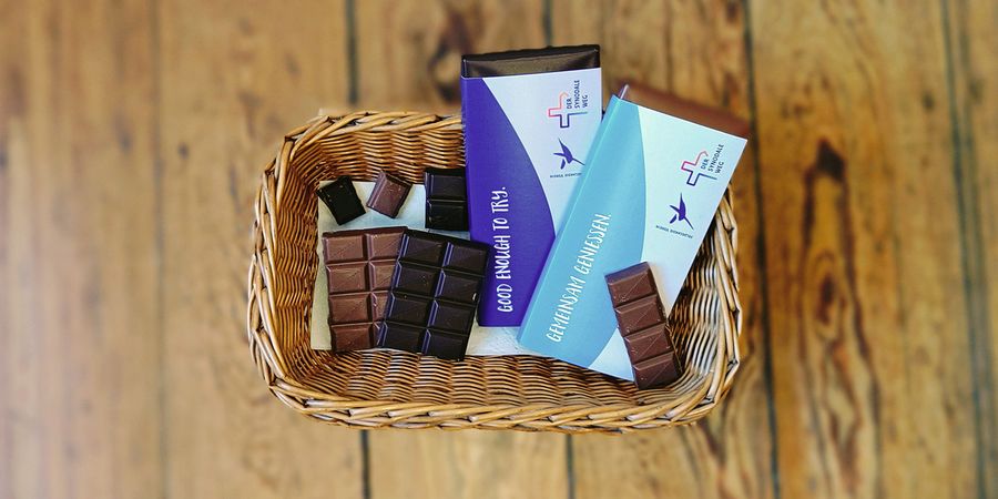 Das Bild zeigt die Sicht auf einen Korb mit Schokolade. Um die Schokolade ist eine Banderole gelegt, die die Logos vom Hildegardis-Verein und vom Synodalen Weg trägt. Mit den Schriftzügen "Good enough to try." (dt. "Gut genug, um es zu probieren") und "Gemeinsam geniessen."